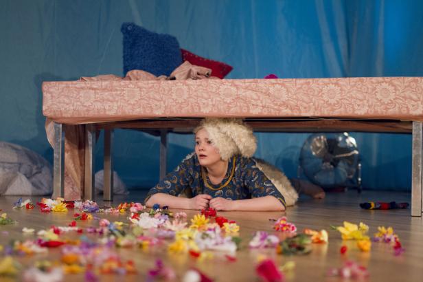 Eine junge Frau liegt unter einem Bett, umgeben von Blumen auf dem Boden.