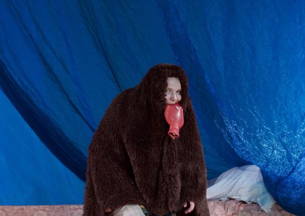 Eine Person in einem braunen Fellmantel hält eine rote Wärmflasche vor dem Mund.