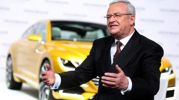 Ein Mann in Anzug gestikuliert vor einem gelben Auto.