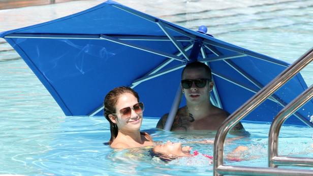 Drei Personen, darunter eine Frau mit Sonnenbrille, entspannen sich in einem Schwimmbad unter einem blauen Sonnenschirm.