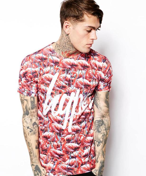 Ein tätowierter Mann trägt ein T-Shirt mit Flamingos und dem Schriftzug „Hype“.