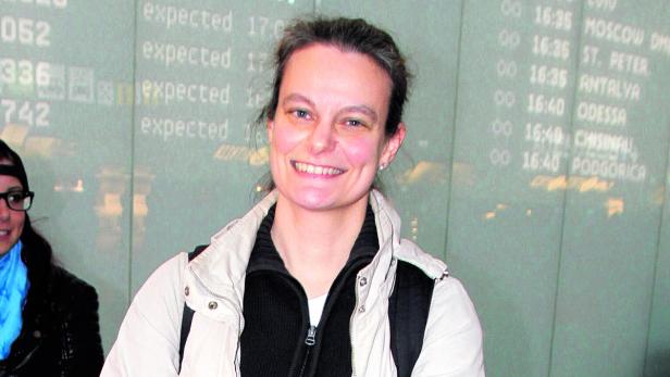 Eine Frau lächelt vor einer Anzeigetafel mit Flugzeiten am Flughafen.