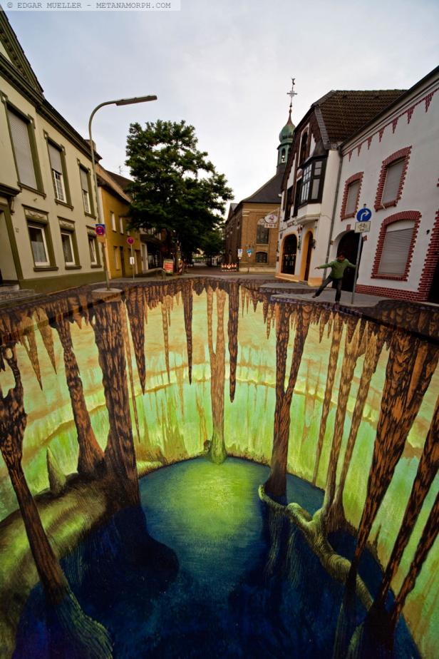 Ein 3D-Straßenkunstwerk zeigt eine scheinbare Höhle in einer Stadt, in der eine Person balanciert.