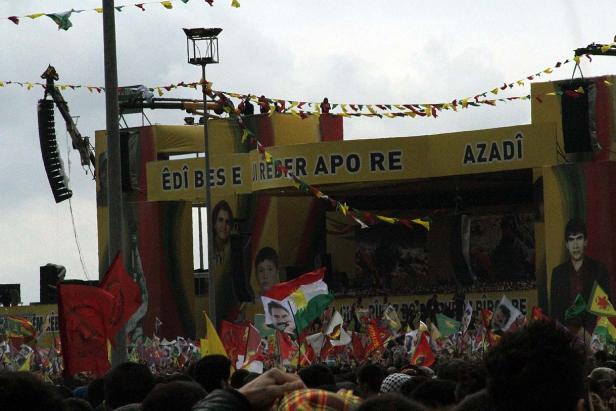 Eine Menschenmenge demonstriert mit kurdischen Flaggen vor einer Bühne mit kurdischen Slogans.