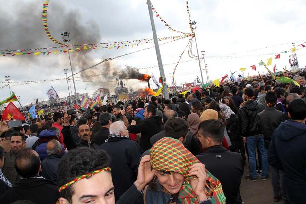 Eine Menschenmenge feiert Newroz mit Fahnen und einem Feuer im Hintergrund.