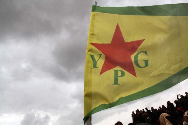 Eine YPG-Flagge weht vor einem bewölkten Himmel über einer Menschenmenge.