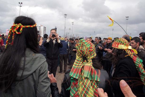 Eine Menschenmenge, einige mit traditionellen kurdischen Kopfbedeckungen, versammelt sich im Freien.