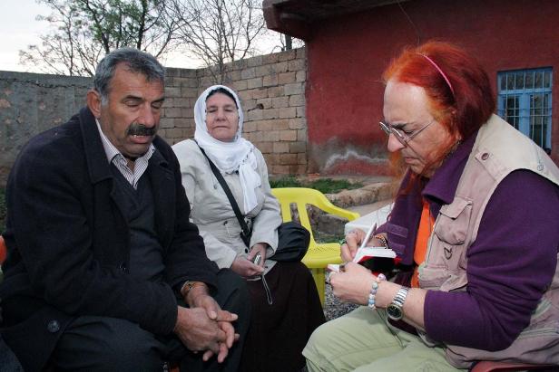 Eine Frau mit roten Haaren schreibt in ein Notizbuch, während zwei Personen daneben sitzen.