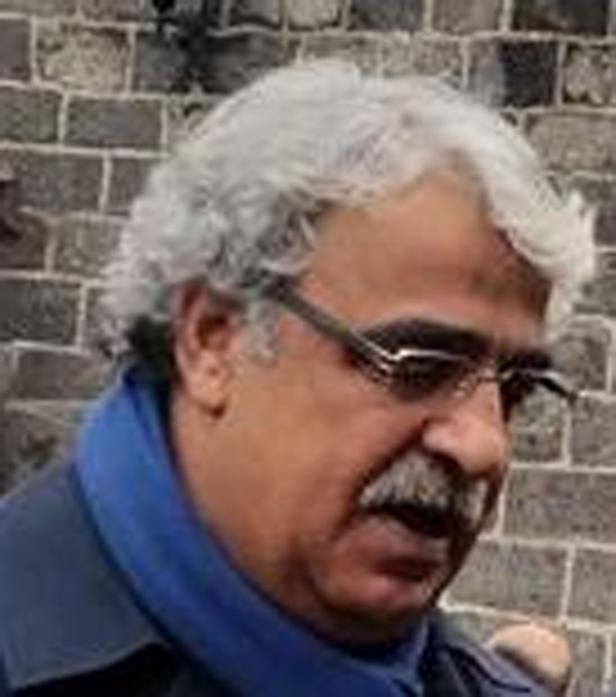 Ein Mann mit Brille, Schnurrbart und grauem Haar vor einer Ziegelwand.