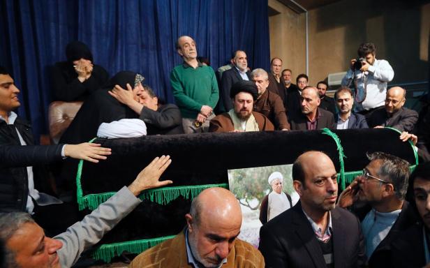 Trauernde versammeln sich um einen Sarg, auf dem ein Porträt eines Geistlichen steht.