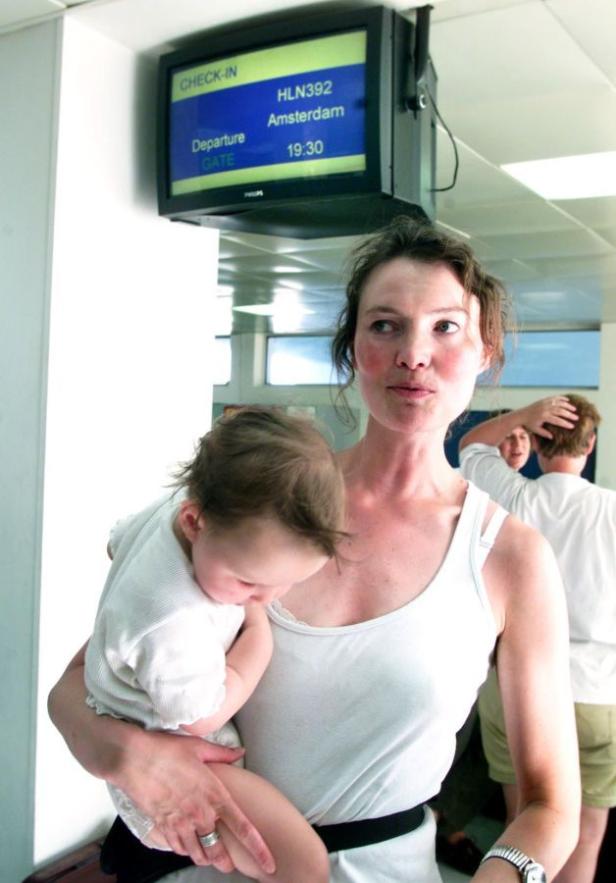 Eine Frau mit einem Baby auf dem Arm steht vor einer Anzeigetafel für den Flug nach Amsterdam.