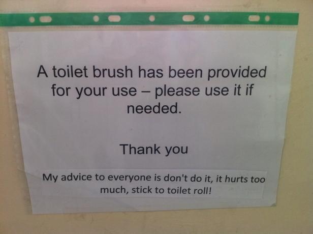 Ein Schild weist auf eine Toilettenbürste hin und gibt humorvolle Ratschläge zur Toilettenpapierverwendung.