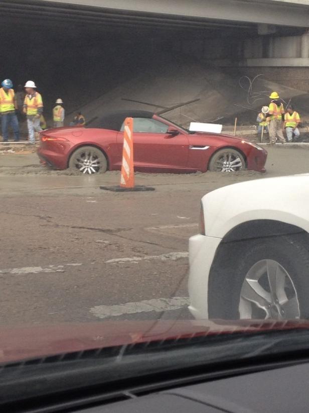 Ein roter Jaguar-Sportwagen steckt im nassen Zement unter einer Brücke fest. Bauarbeiter stehen daneben.
