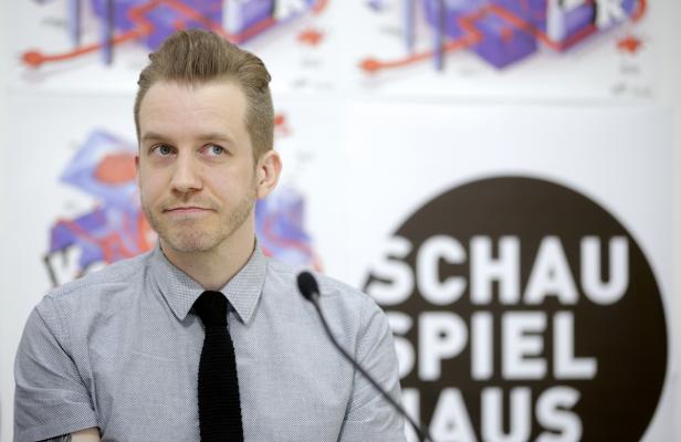 Ein Mann mit Hemd und Krawatte steht vor einem „Schauspielhaus“-Logo.