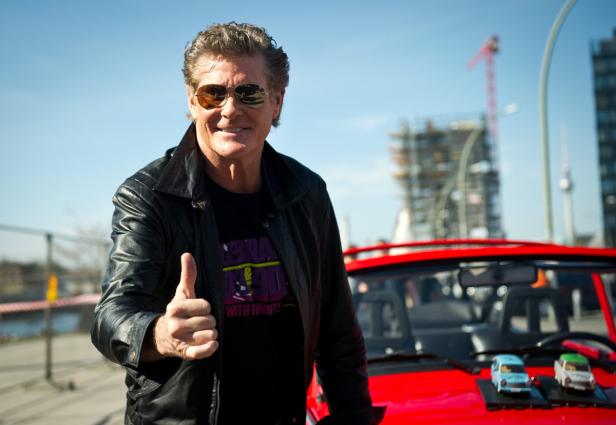 David Hasselhoff posiert vor einem roten Auto mit Miniatur-Trabis auf der Motorhaube.