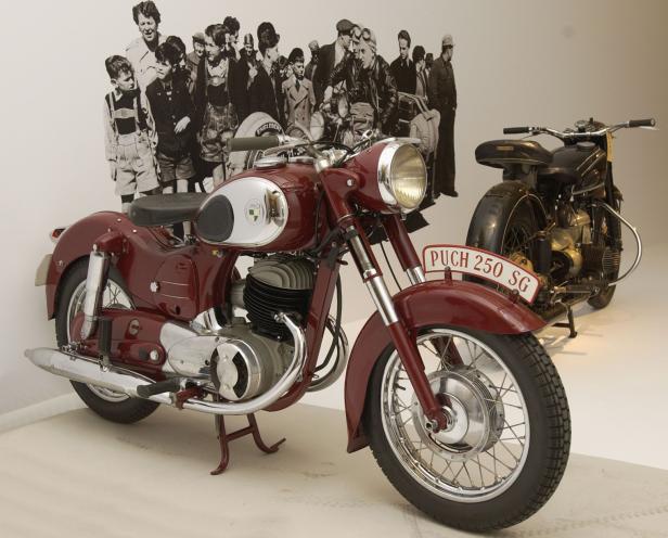 Eine rote Puch 250 SG steht vor einer Fotowand mit Menschen.