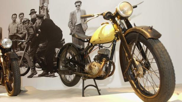 Ein gelbliches Motorrad steht vor einer Fotowand mit historischen Aufnahmen.