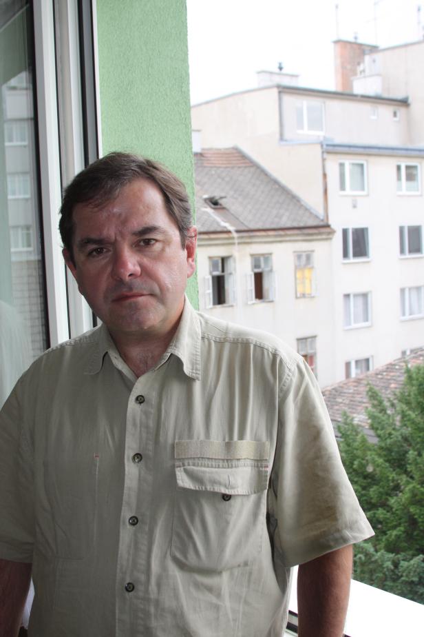 Ein Mann in einem hellen Hemd steht vor einem Fenster mit Blick auf ein Wohngebiet.