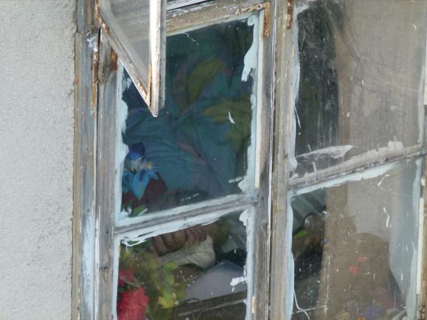 Ein zerbrochenes Fenster mit Blumen und Stoffresten dahinter.