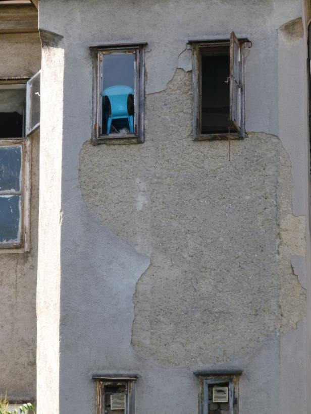 Ein blau Stuhl steht hinter einem Fenster in einem baufälligen Gebäude.