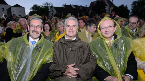 Drei Männer sitzen in der ersten Reihe einer Veranstaltung, viele tragen gelbe Ponchos.