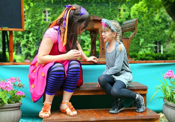 Zwei Mädchen in Kostümen sitzen auf einer Bühne vor einer grünen Wand.