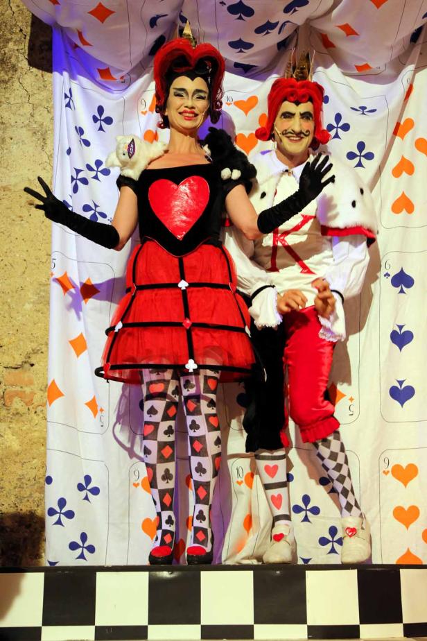 Eine als Herzkönigin und Herzkönig verkleidete Frau und ein Mann posieren vor einem Vorhang mit Spielkartensymbolen.