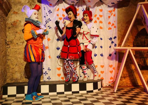 Drei Darsteller in Kostümen, die an „Alice im Wunderland“ erinnern, stehen auf einer Bühne.