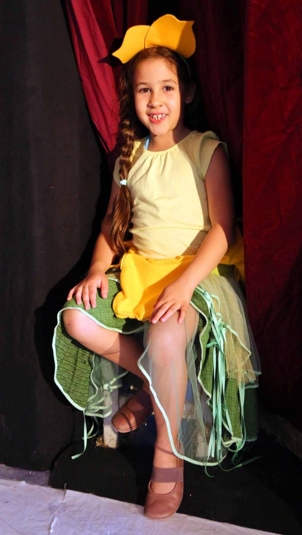 Ein junges Mädchen in einem gelben und grünen Blumenkostüm sitzt auf einer Bühne.