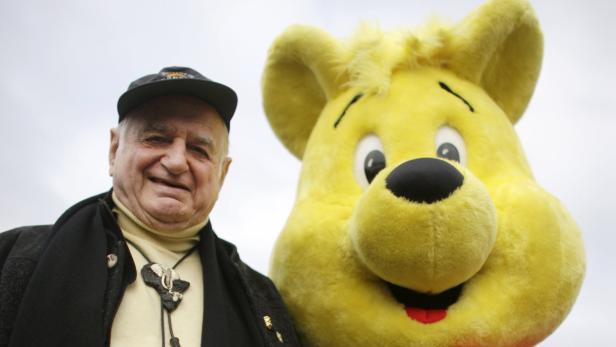 Ein älterer Mann mit Kappe posiert neben einer großen gelben Plüschfigur.