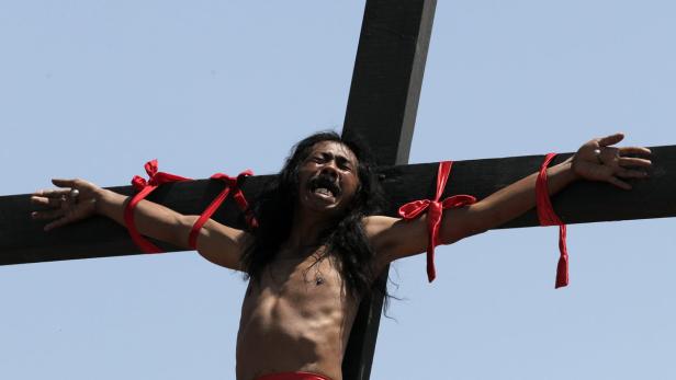 Ein Mann ist mit roten Bändern an ein Holzkreuz gebunden.