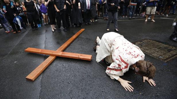 Eine Frau kniet auf der Straße neben einem Holzkreuz, ihr Gewand ist mit roter Farbe befleckt.
