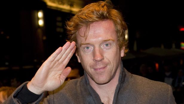 Damian Lewis grüßt mit erhobener Hand.