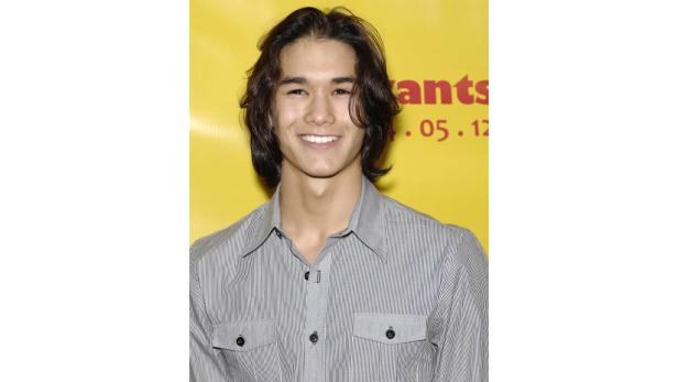 Ein lächelnder junger Mann mit langen, dunklen Haaren vor einem gelben Hintergrund.