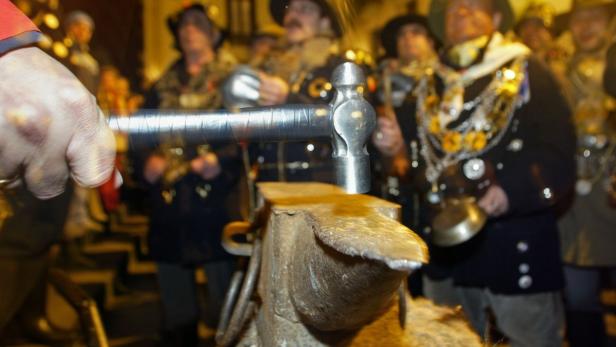 Eine Person hämmert auf einem Amboss, umgeben von Menschen in Trachten.