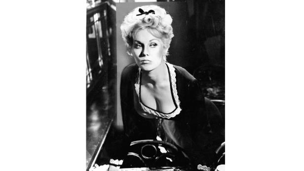 Eine Frau mit blonden, hochgesteckten Haaren und dunklem Kleid blickt zur Seite.