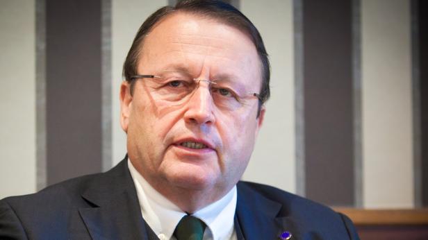 Ein Mann mit Brille und dunklem Anzug vor einem gestreiften Hintergrund.