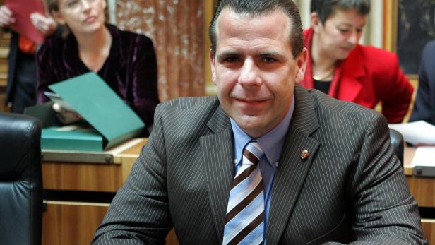 Ein Mann im Anzug sitzt an einem Tisch mit anderen Personen im Hintergrund.