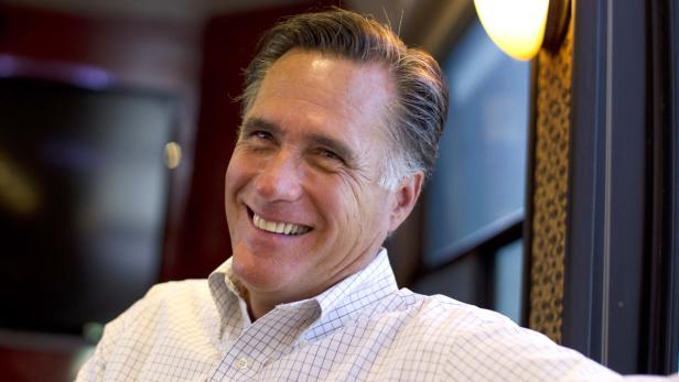 Ein lächelnder Mann in einem hellen Hemd sitzt in einem Raum mit einem Fernseher im Hintergrund.