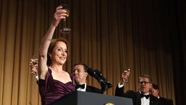 Eine Frau in einem lila Kleid hält ein Weinglas in die Höhe, während sie an einem Rednerpult steht.