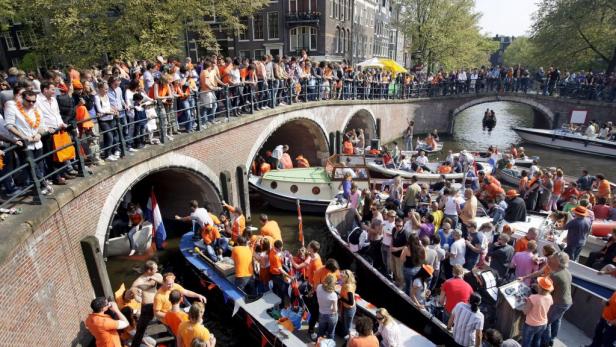 Menschen feiern auf Booten und Brücken in Amsterdam, viele tragen orange Kleidung.
