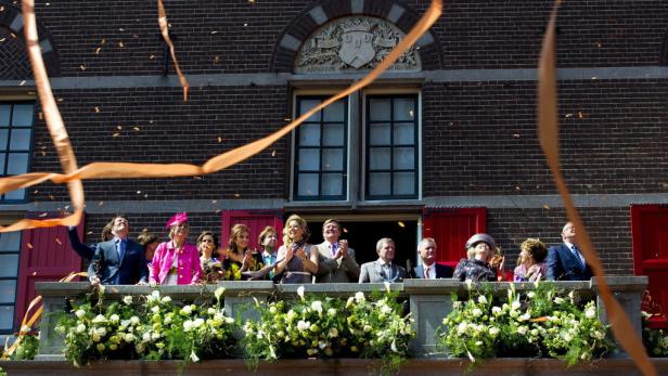 König Willem-Alexander und Königin Máxima winken von einem Balkon, während orangefarbene Bänder in der Luft schweben.