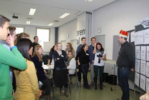 Eine Gruppe junger Leute steht in einem Raum und diskutiert mit einem Mann mit Krone vor einer Pinnwand.