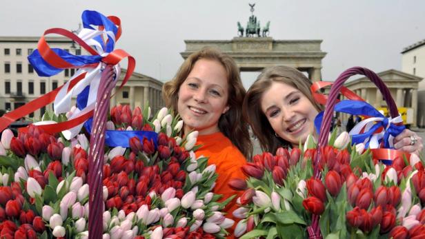 Zwei Frauen lächeln hinter Körben voller Tulpen vor dem Brandenburger Tor.