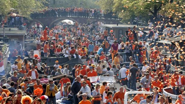 Menschen in orangefarbener Kleidung feiern auf Booten und einer Brücke in Amsterdam.