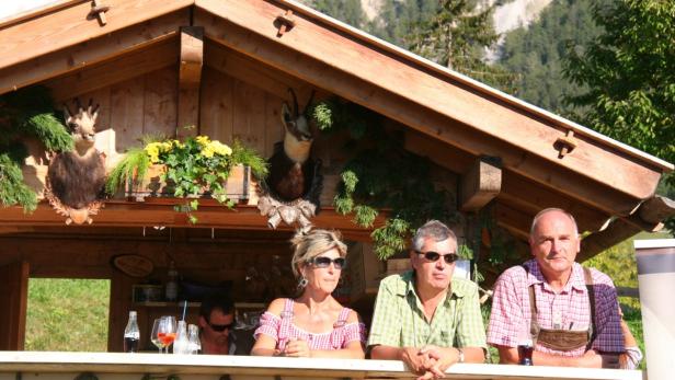 Drei Personen stehen an einer Bar im Freien mit Bergkulisse.