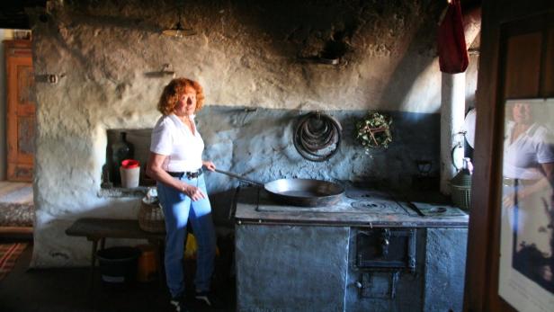 Eine Frau steht in einer rustikalen Küche und hantiert mit einer großen Pfanne auf einem alten Herd.