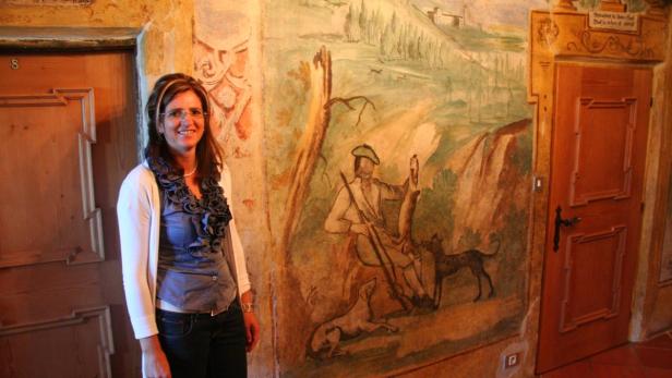 Eine Frau steht vor einer Wandmalerei mit einer Jagdszene.