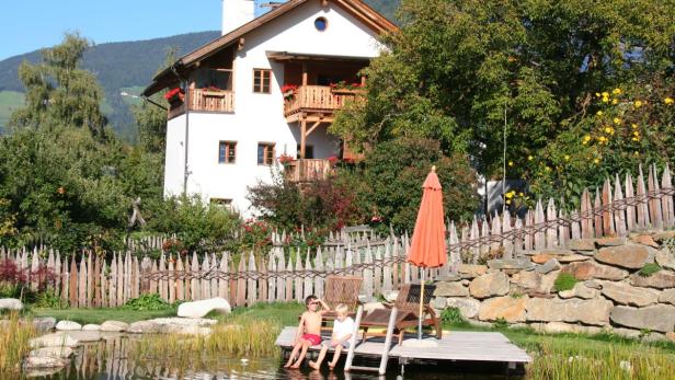 Zwei Kinder sitzen auf einem Holzsteg an einem Teich vor einem Haus.