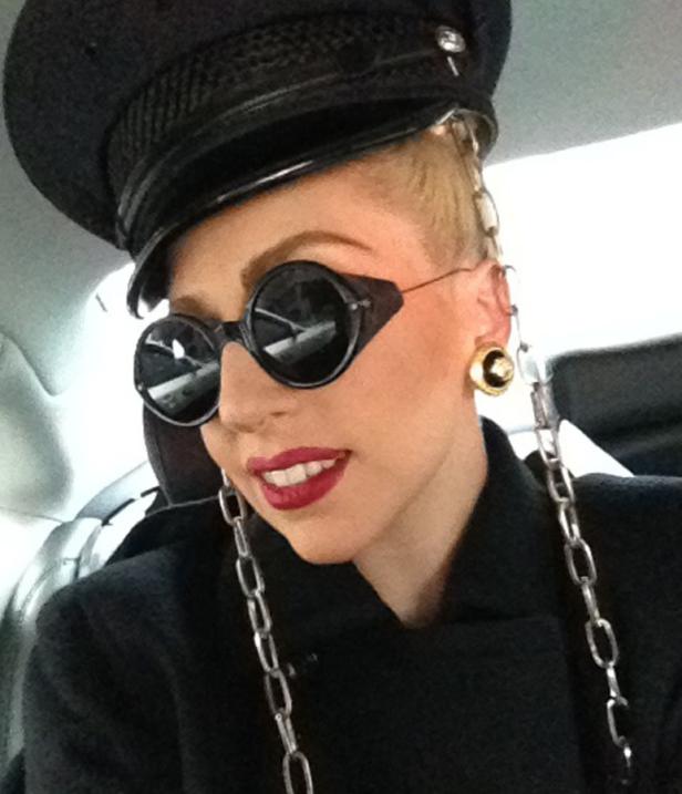 Lady Gaga mit runden Brillengläsern, einer Kapitänsmütze und Ketten als Ohrringe.